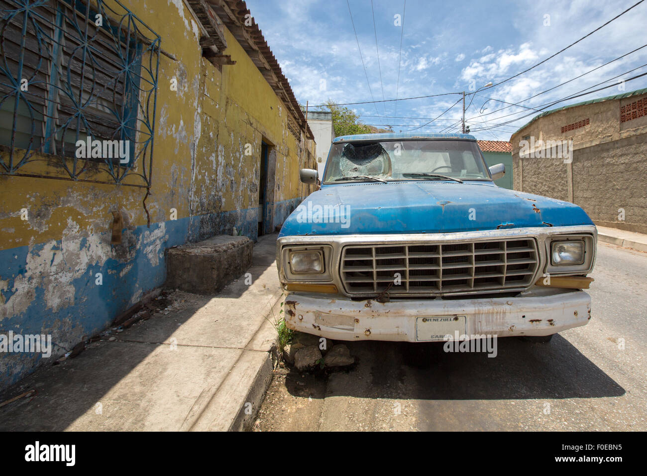 Rusted blue vintage car with smashed window parked in dirty street of Margarita Island against a blue sky. Venezuela Stock Photo