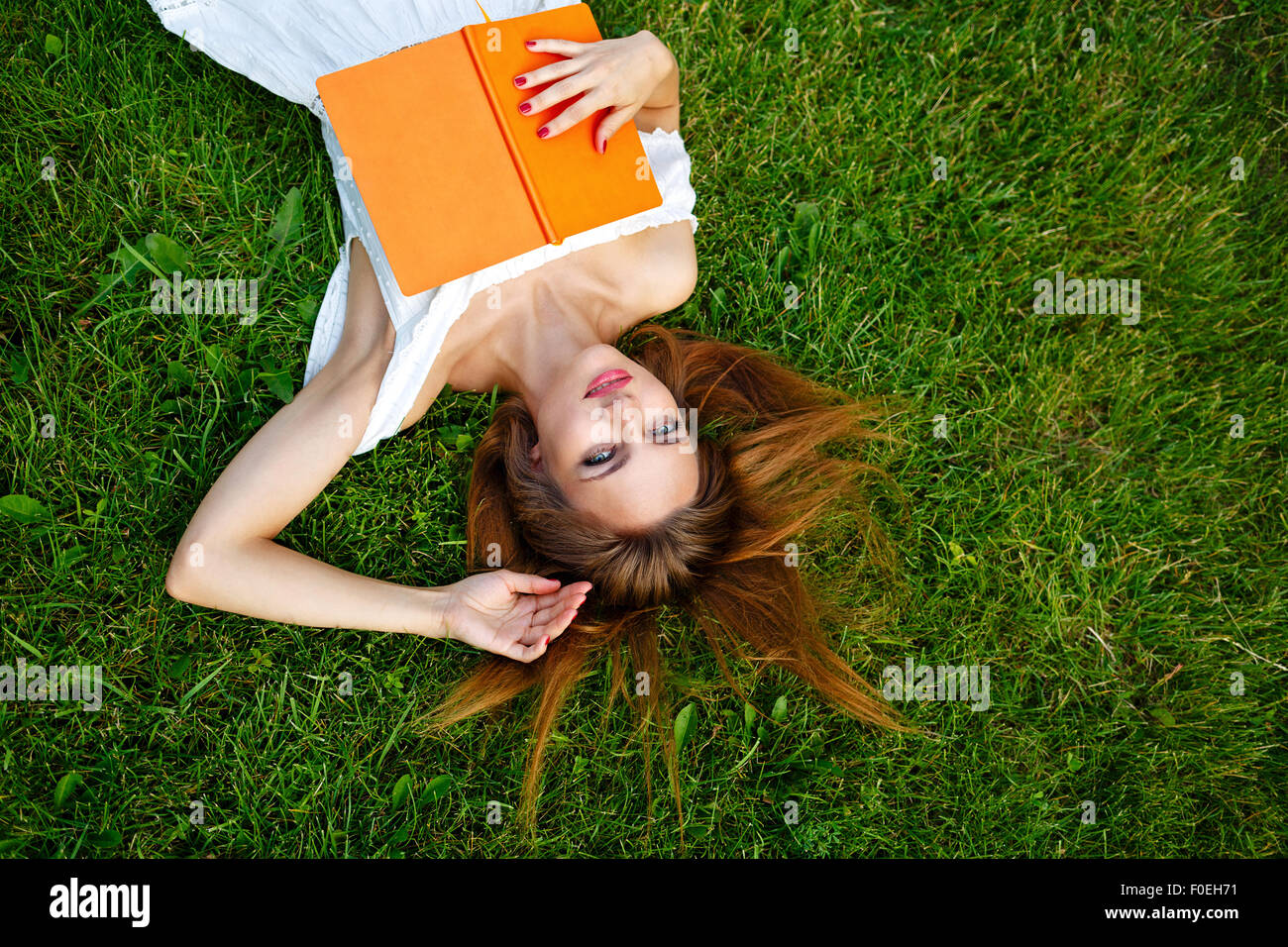 A young woman relaxes with a book lying on a green lawn in the park ...