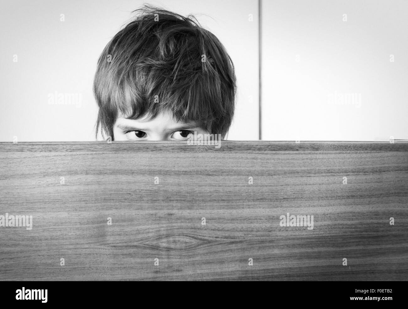 Scared little girl hiding. She is hiding behind a wooden surface, peeking with a worried look on her face. Stock Photo
