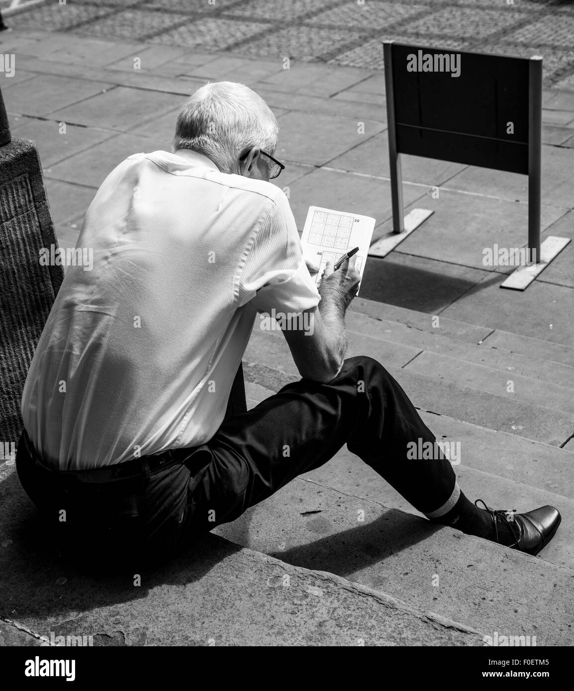 Photo of an elderly man sitting on steps and doing a crossword or sudoku puzzle Stock Photo