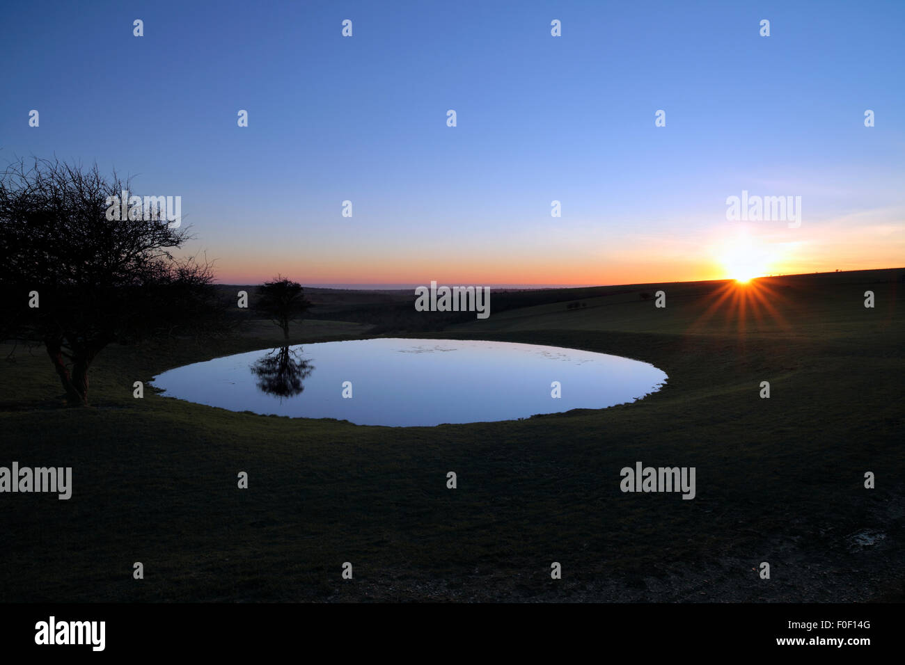 The sun setting behind a dew pond on the South Downs, near Ditchling Beacon, East Sussex. Stock Photo