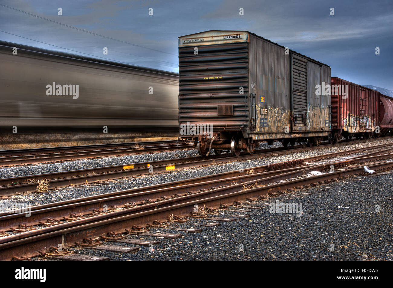 A fast moving freight train is blurred behind a static box car on the fore ground train track Stock Photo
