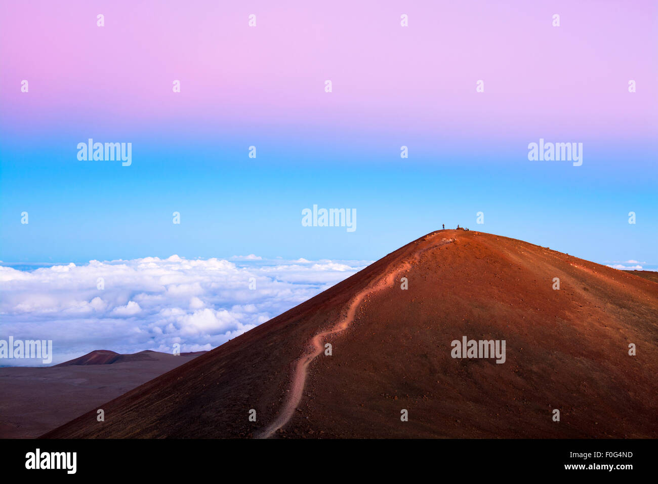 A red dirt mountain with a trail to the top at 14,000 feet overlooks the top of clouds and exposes the pink inversion layer Stock Photo