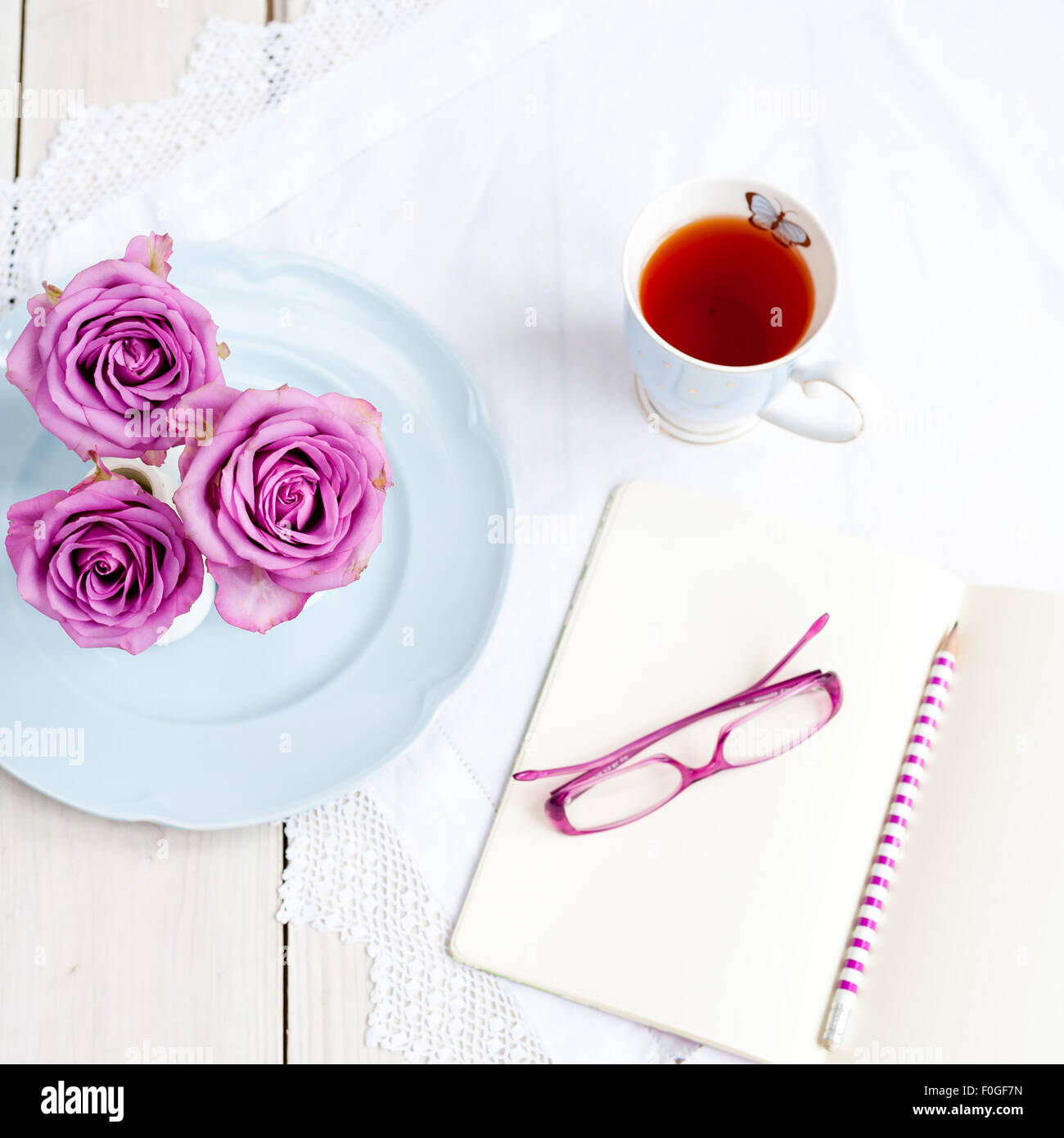 three pink roses in vases on blue plate with mug of tea and open notebook with glasses and pencil Stock Photo