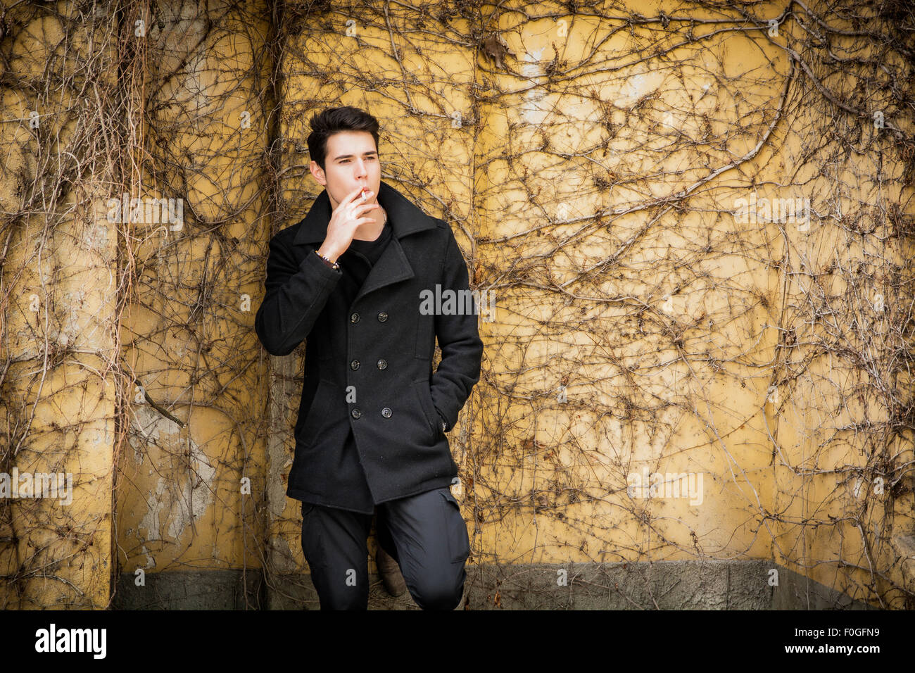 Handsome serious young man standing against plant covered wall, wearing winter clothes, smoking cigarette Stock Photo