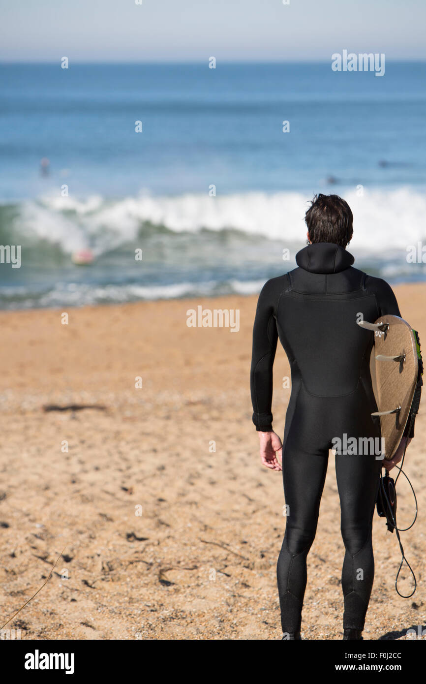 Young attractive surfer man standing on a beach and carrying his surfing board while looking at the Atlantic Ocean during a sunn Stock Photo