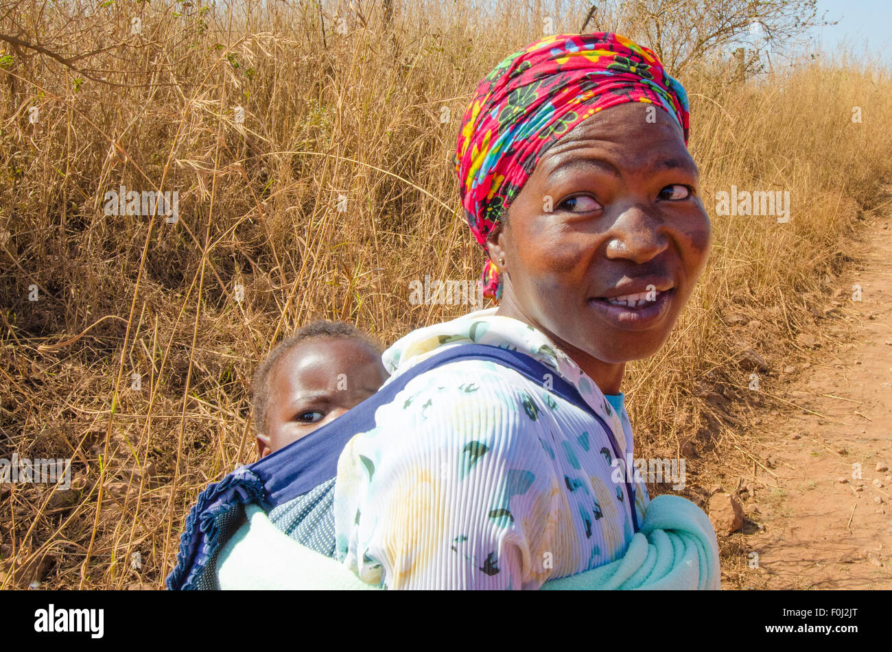 A mother carries her young son on her back as she hikes through the dirt roads of rural Swaziland. Stock Photo