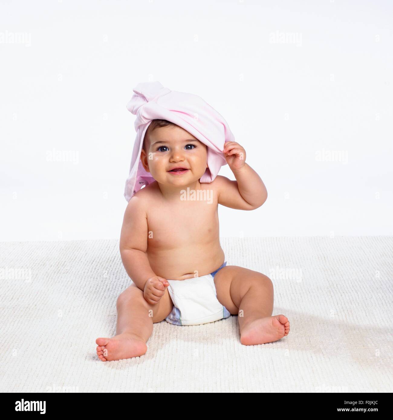 Baby boy wearing nappy, with pink shirt on head, looking at camera