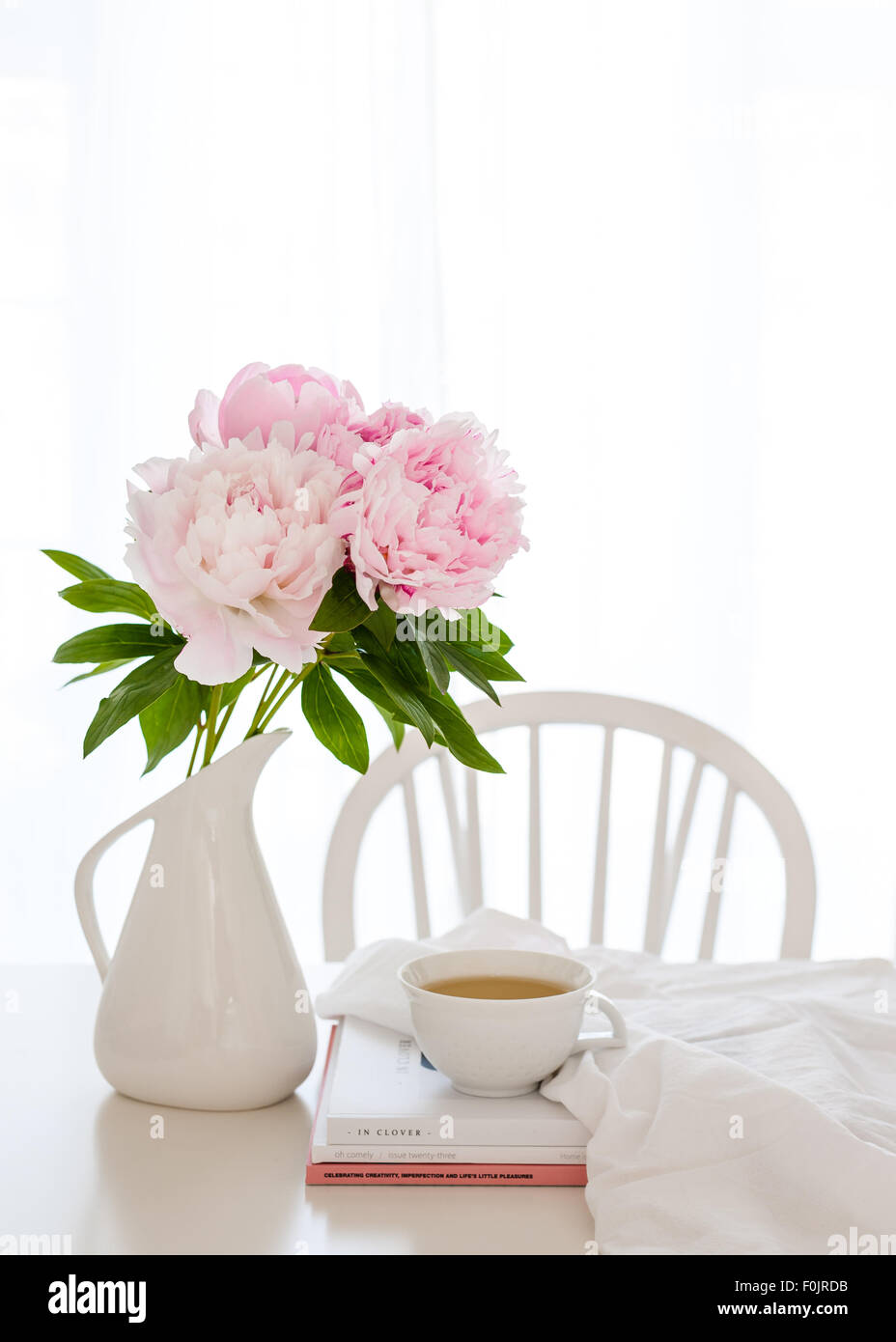 a bunch of pink peonies in a white jug on the table, with magazines, cup of tea, fabric and chair in the background Stock Photo