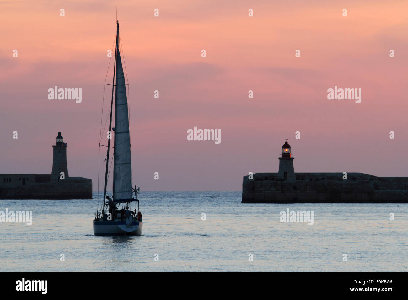 Sailing yacht departing from Malta's Grand Harbour at dawn, with breakwater lighthouses visible Stock Photo