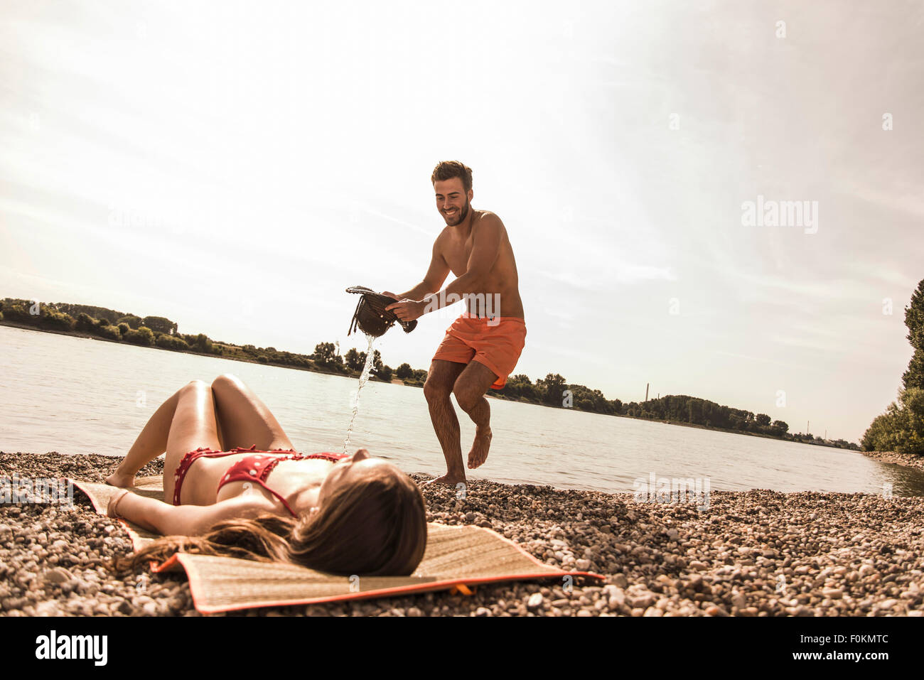 Playful young man trying to pour water over girlfriend sunbathing by the riverside Stock Photo