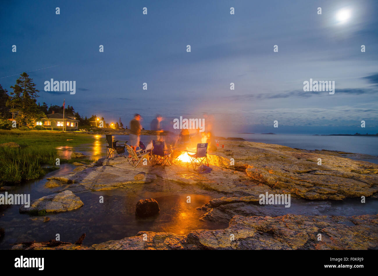 Family and friends warm up by a bonfire under a moon-lit night as high tide slowly trickles inland. Stock Photo