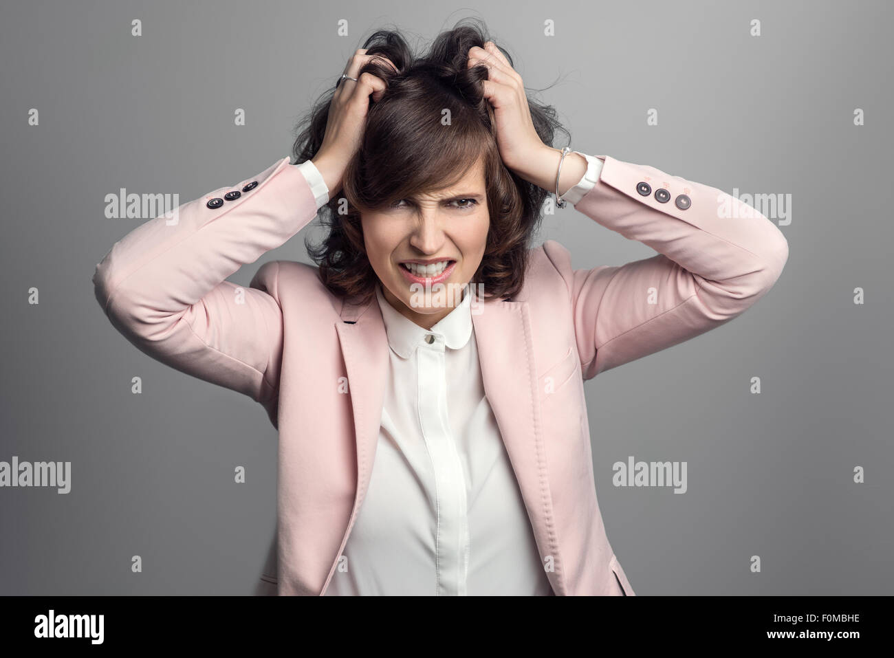 Attractive stylish young woman in a pink jacket tearing at her brown hair with her hands as she vents her frustration, over grey Stock Photo