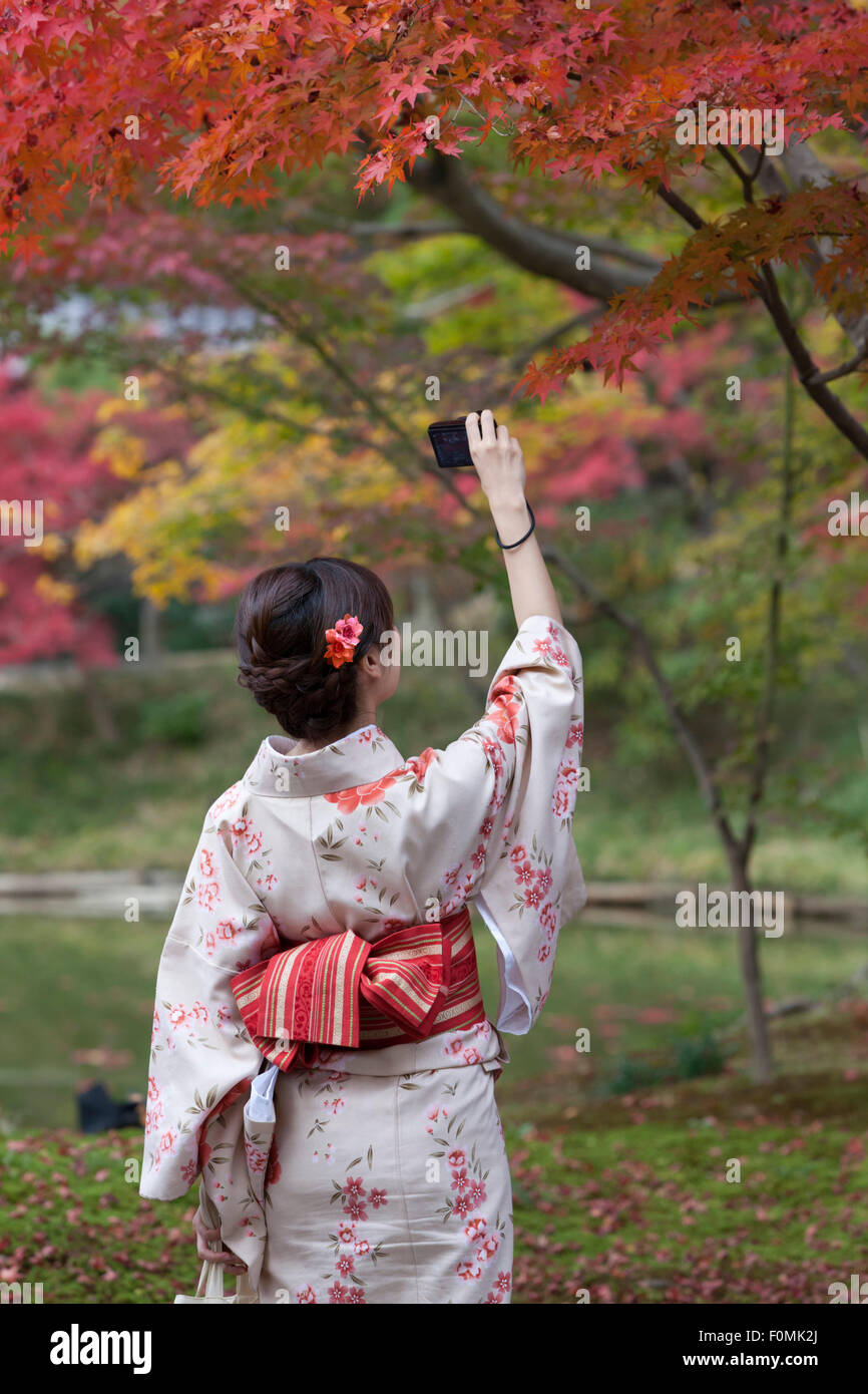 Young Japanese girl in traditional kimono photographing brilliant autumn maple leaves showcasing Kyoto fall foliage photography opportunities
