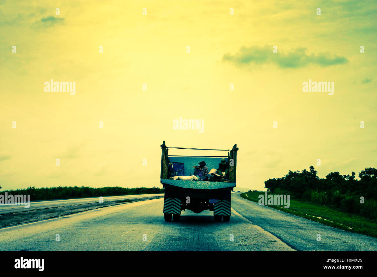 Two people traveling in back of a truck on an empty road in Cuba. The picture has a toned effect and very contrasted. Stock Photo