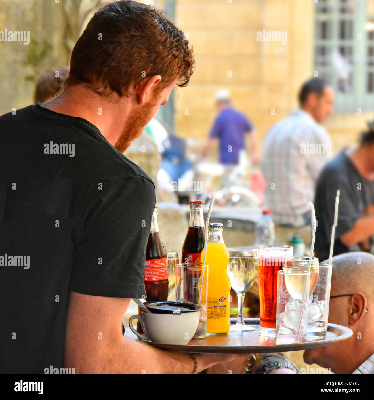 French waiter carries tray full of drinks to customers at outdoor bar tables in Place de l'Hotel