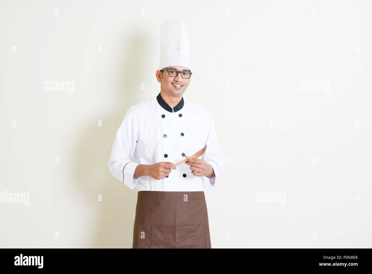 Portrait of handsome Indian male chef in uniform hand holding spatula and smiling confidently, standing on plain background with Stock Photo