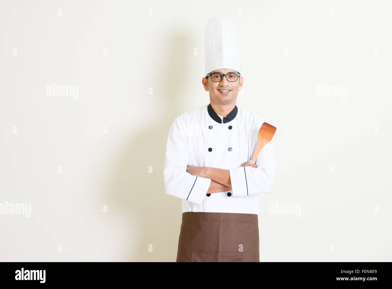 Portrait of handsome Indian male chef in uniform hand holding spatula and smiling, standing on plain background with shadow, cop Stock Photo