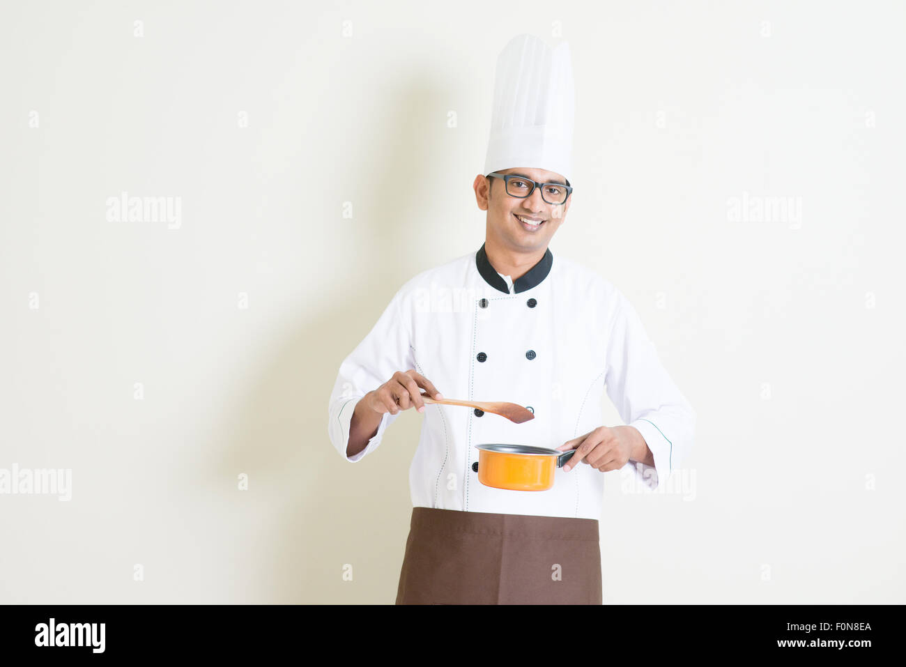 Portrait of handsome Indian male chef in uniform holding cooking pot and stirring food, standing on plain background with shadow Stock Photo