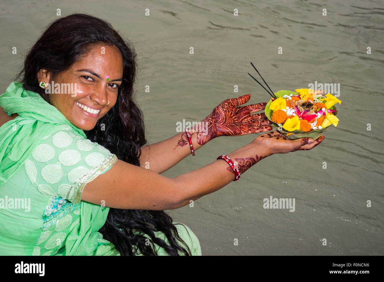 A young woman with long black hair, henna painted hands and a green dress is holding a deepak, a flower offering, at the ghats Stock Photo