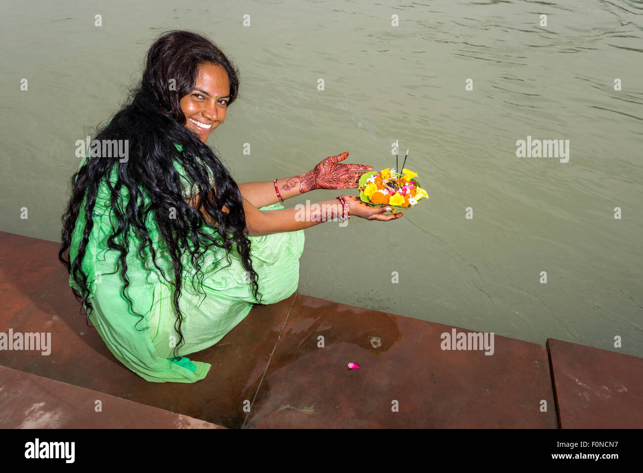 A young woman with long black hair, henna painted hands and a green dress is holding a deepak, a flower offering, at the ghats Stock Photo