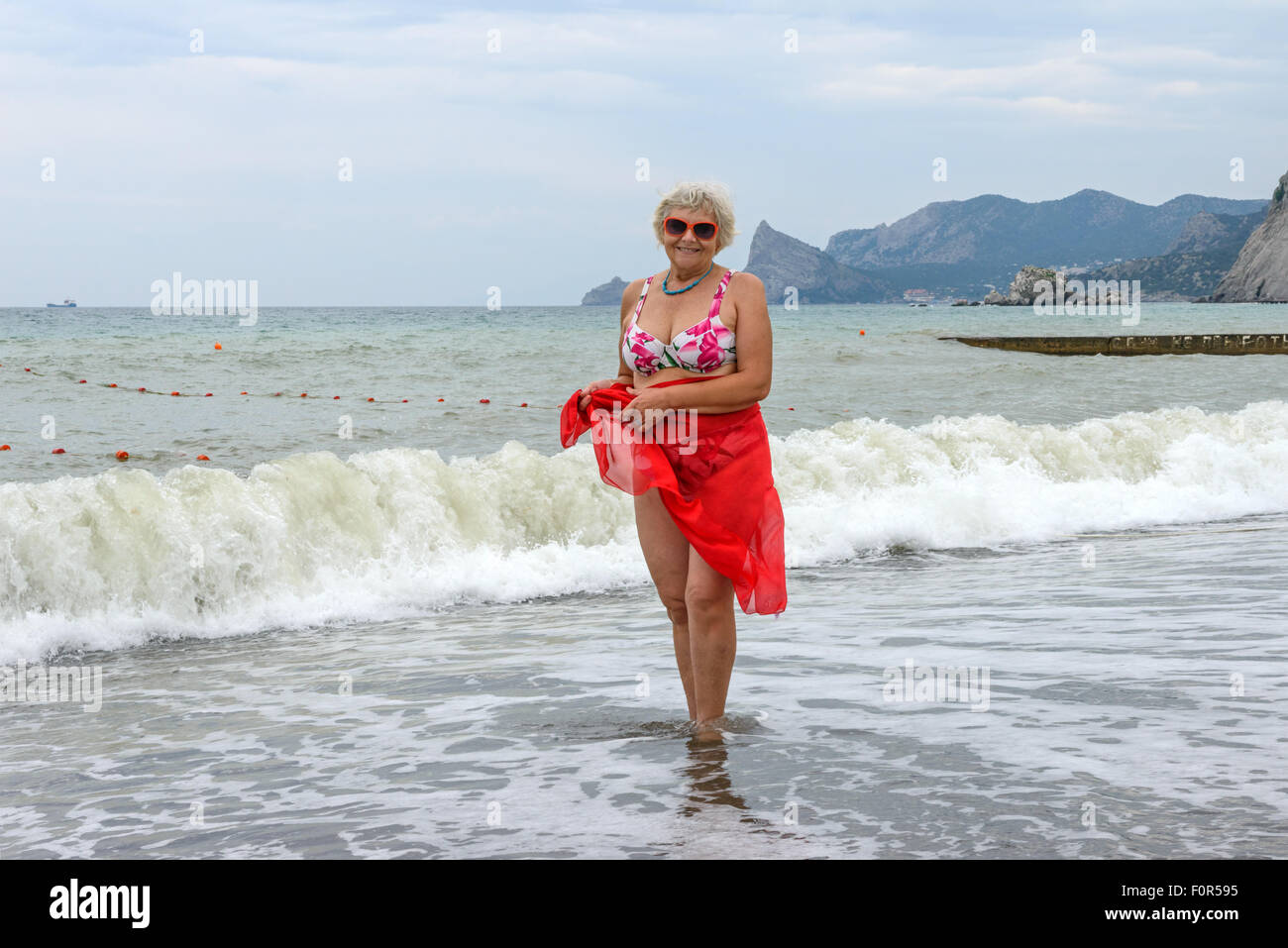 Full length portrait of elderly woman that is standing on pebble beach with beach wrap on her hips on surf waves and cliff backg Stock Photo