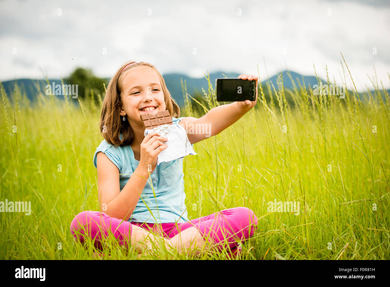 Child is taking photo with mobile phone camera while eating chocolate outdoor in nature Stock Photo