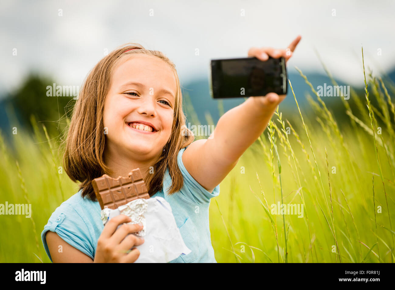 Child is taking photo with mobile phone camera while eating chocolate outdoor in nature Stock Photo