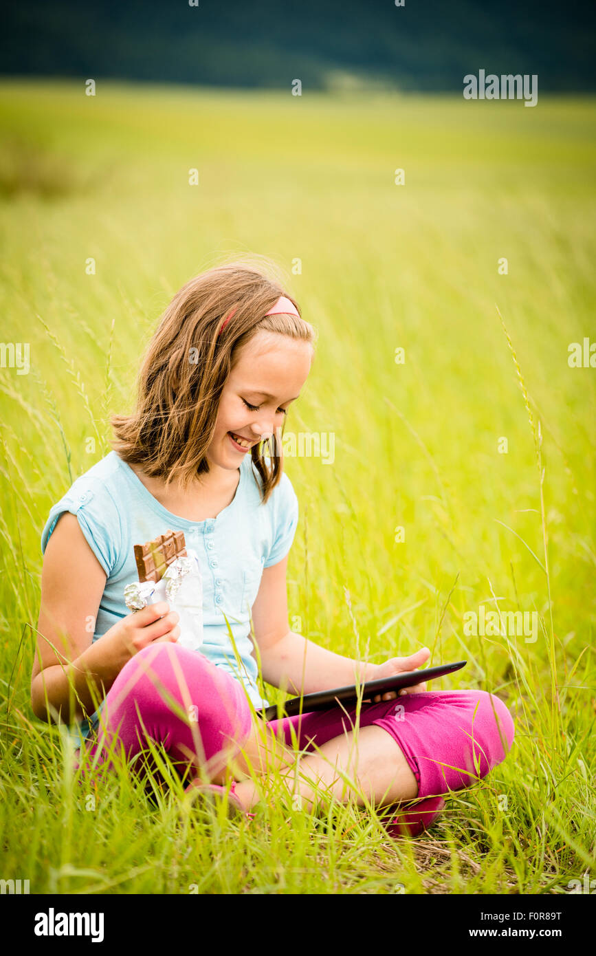 Child eating and relishing chocolate while watching tablet - outdoor in nature Stock Photo