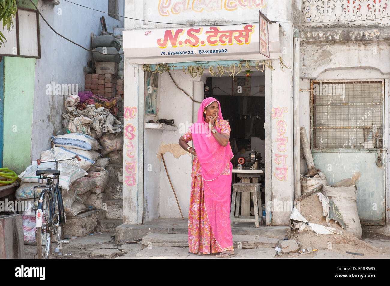 Rajasthan, India. Between Jodhpur and Jaipur. Woman standing outside her tiny sewing shop with her sewing machine in traditional Rajasthan pink sareelooking along the street with her fingers over her mouth. Stock Photo