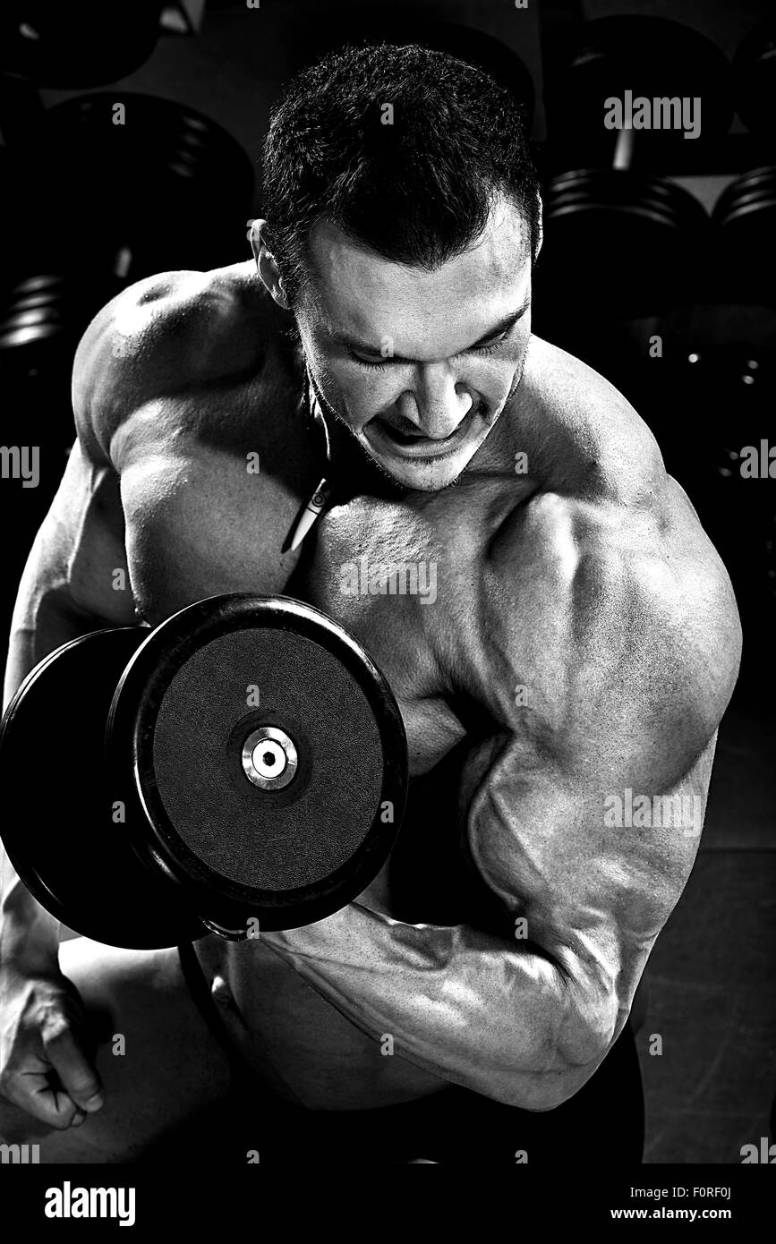 vertical close up portrait handsome guy bodybuilder ,  execute exercise with  dumbbells, in dark gym, balck and white photo Stock Photo