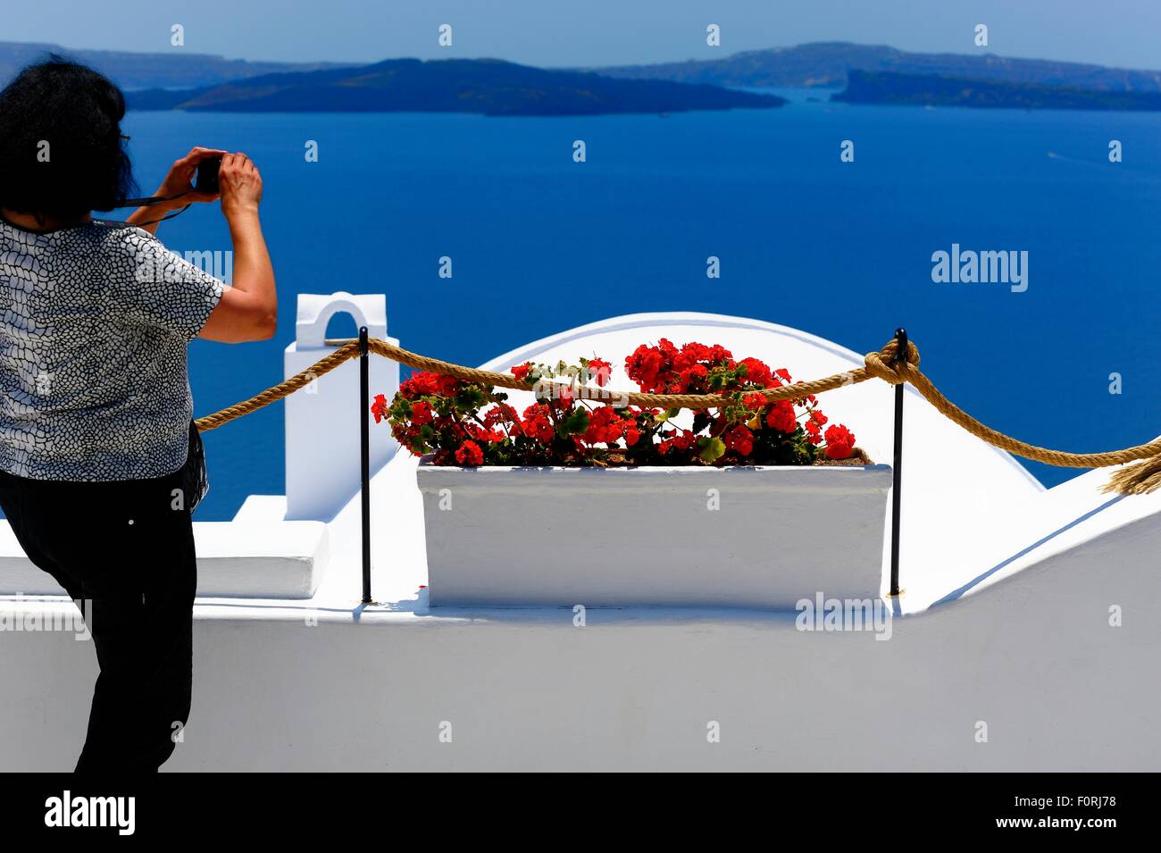 A woman taking a photograph of a red flower box overlooking the caldera in Oia Santorini Greece. Stock Photo