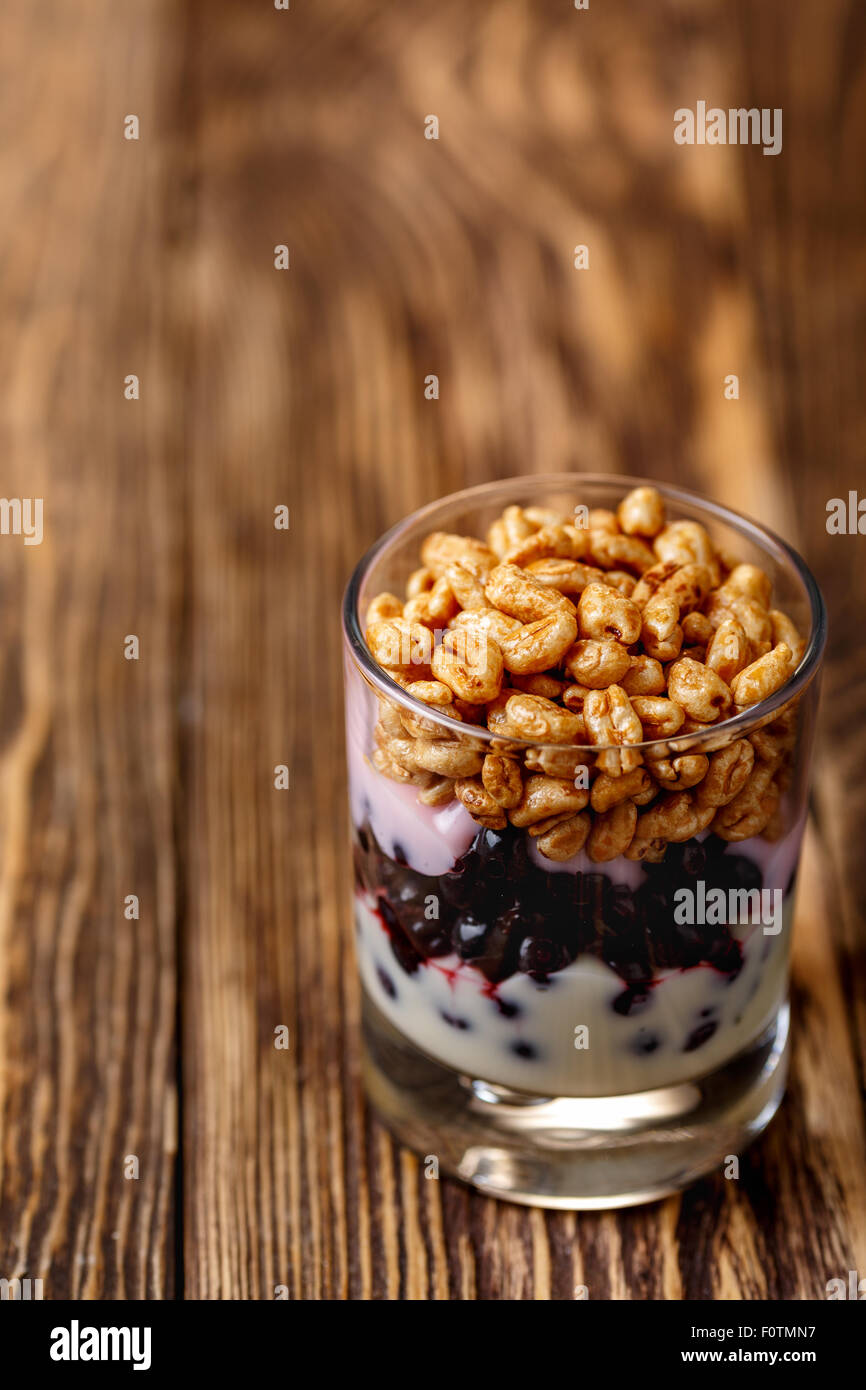 Parfait-style healthy layered snack or dessert with yogurt,bilberry jam,fresh bilberries,puffed honey wheat in  glass on wooden  Stock Photo