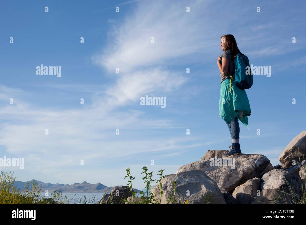 Rear view of young woman wearing backpack standing on rocks looking away, Great Salt Lake, Utah, USA Stock Photo