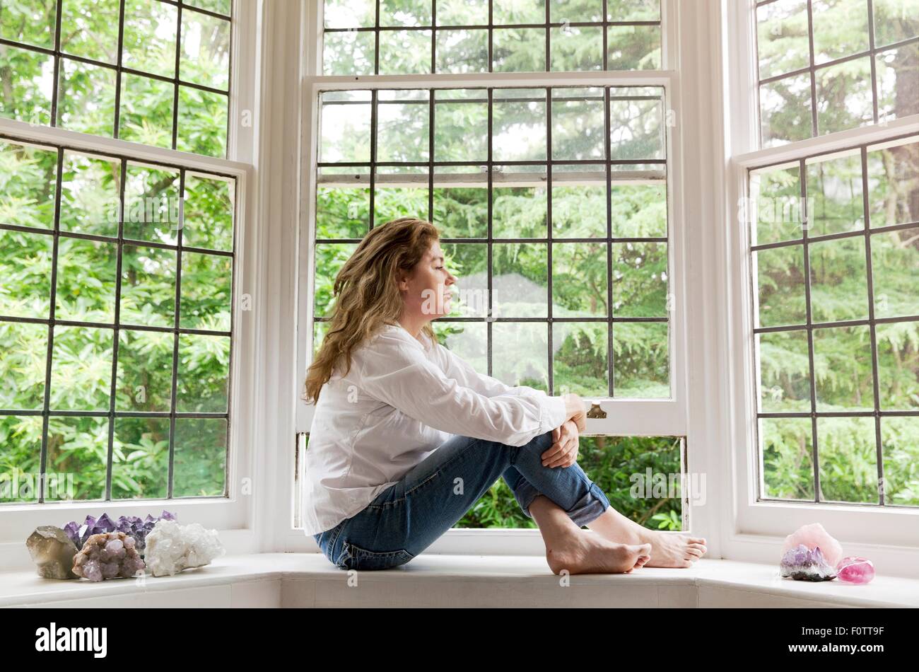 Portrait of mature woman sitting on living room windowsill looking out of window Stock Photo