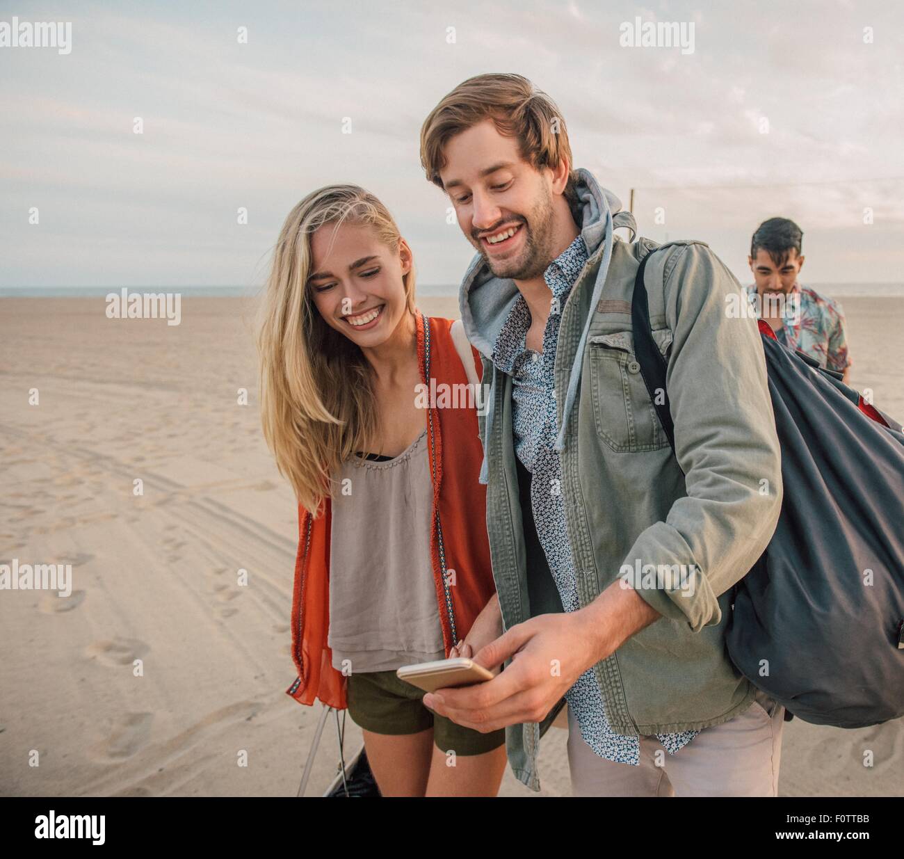 Group of friends walking along beach, young couple looking at smartphone Stock Photo