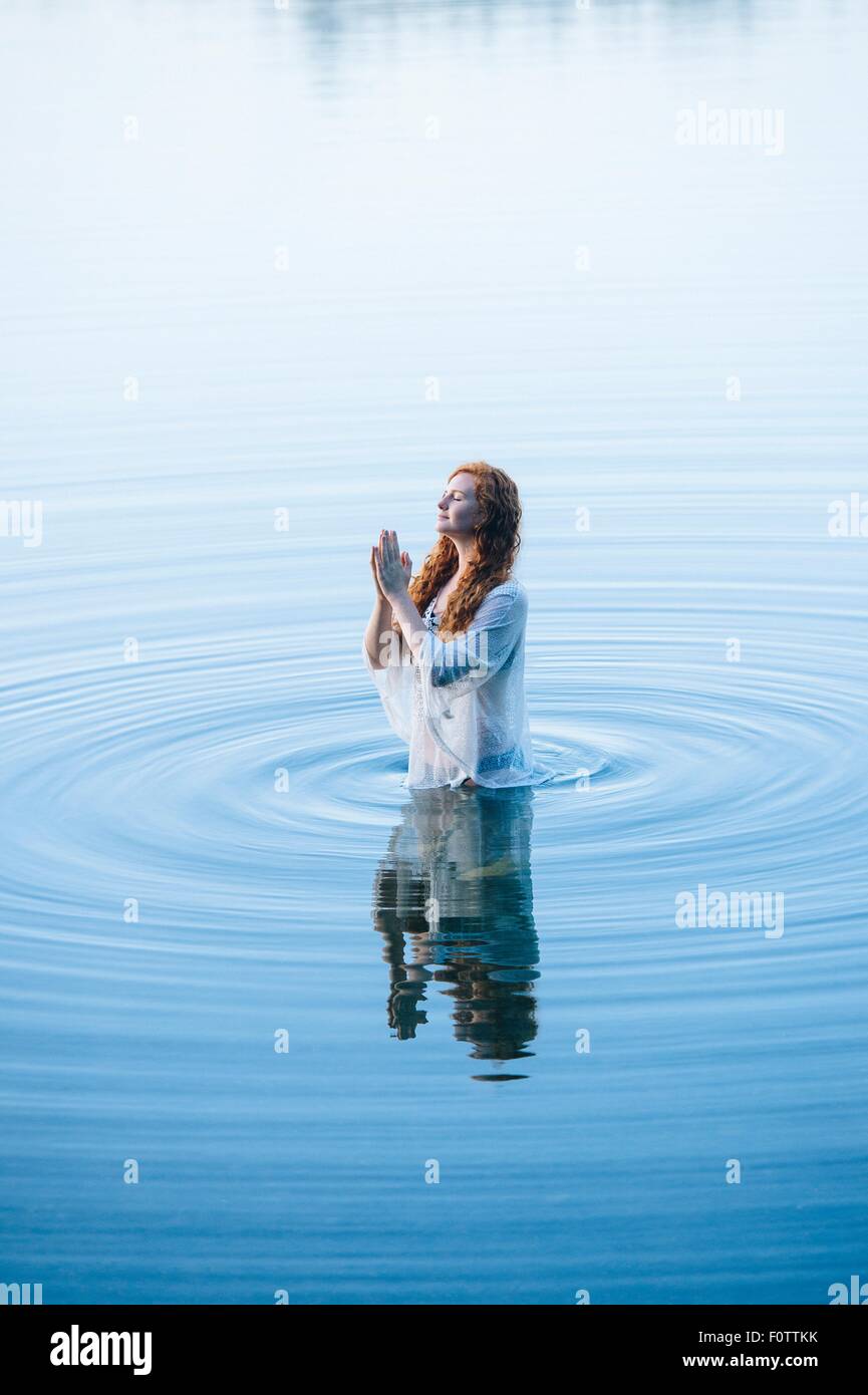 Young woman standing in lake ripples with eyes closed and hands together Stock Photo