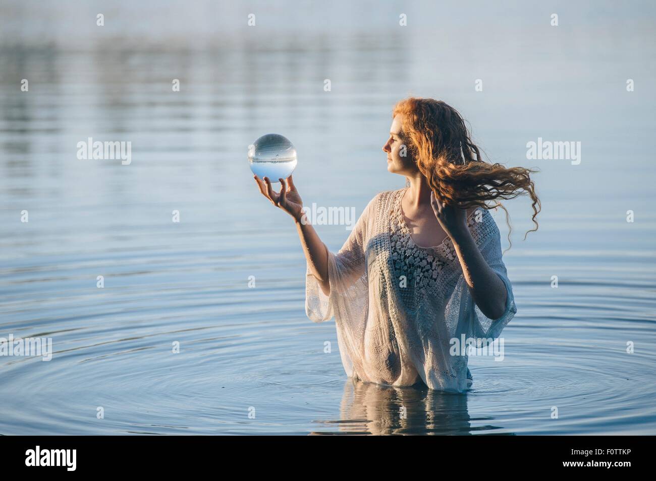 Young woman with long red hair standing in lake gazing at crystal ball Stock Photo