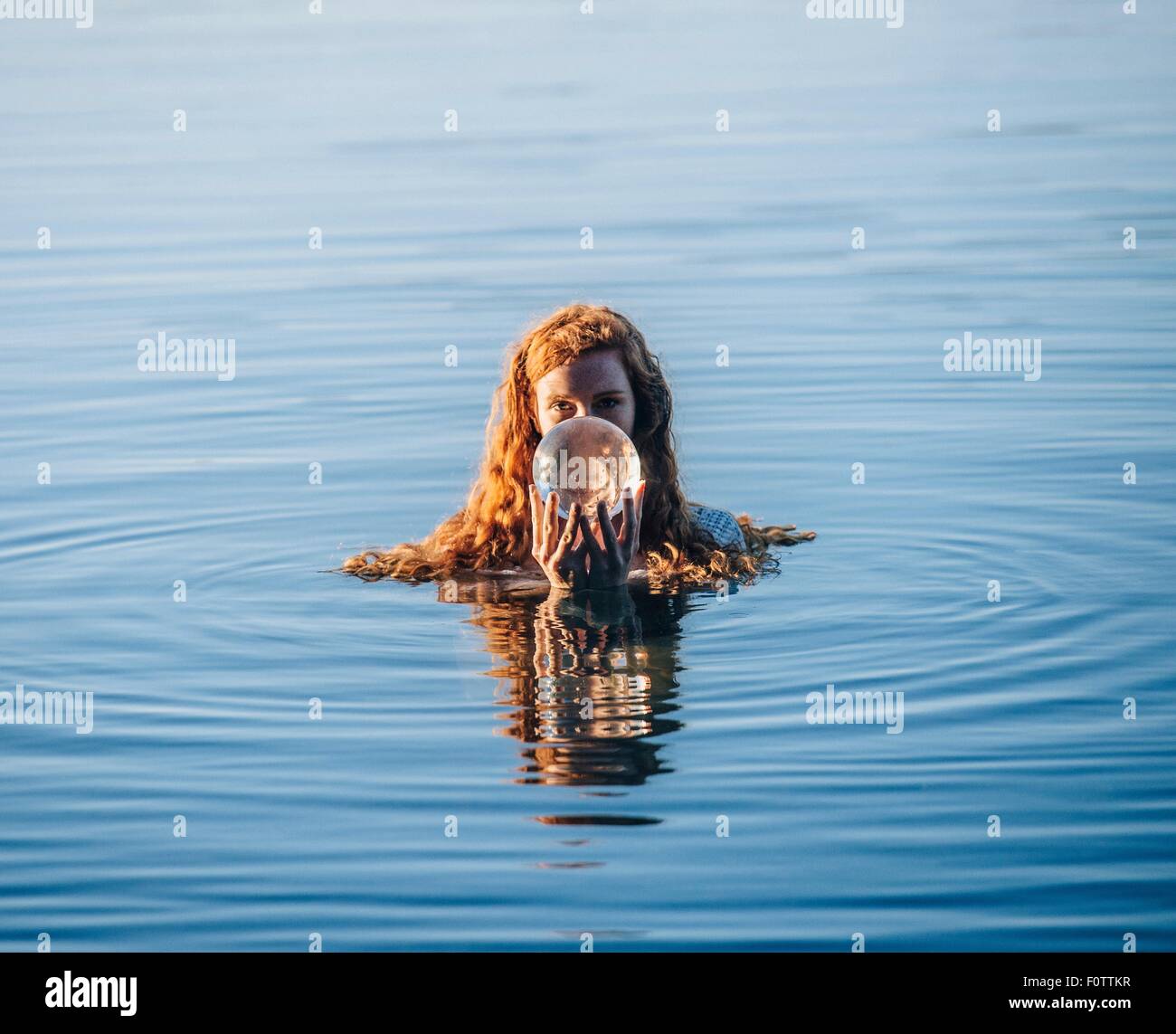 Head and shoulders of young woman with long red hair in lake holding crystal ball Stock Photo