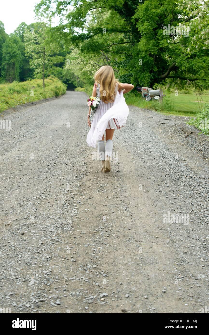 Rear view of young woman walking on rural road with flowers behind her back Stock Photo