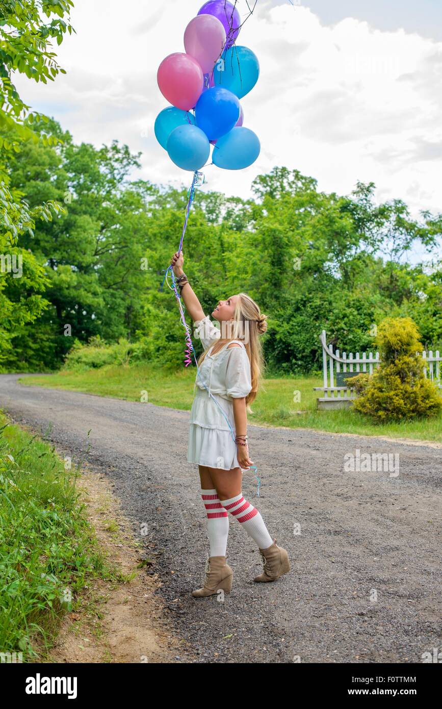 Young woman gazing up at bunch of balloons on rural road Stock Photo