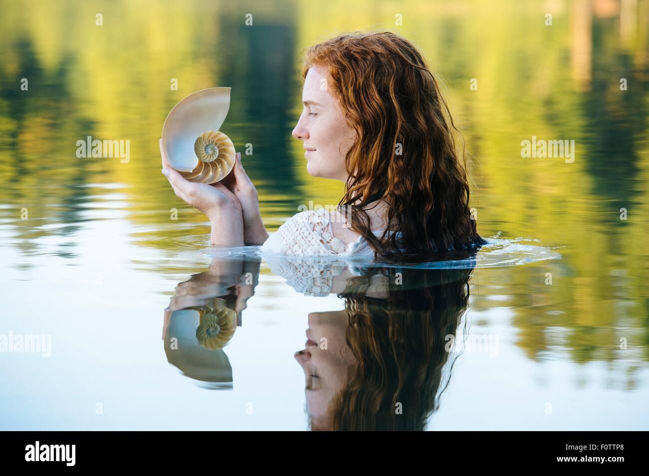 Head and shoulders of young woman with long red hair in lake gazing at seashell Stock Photo