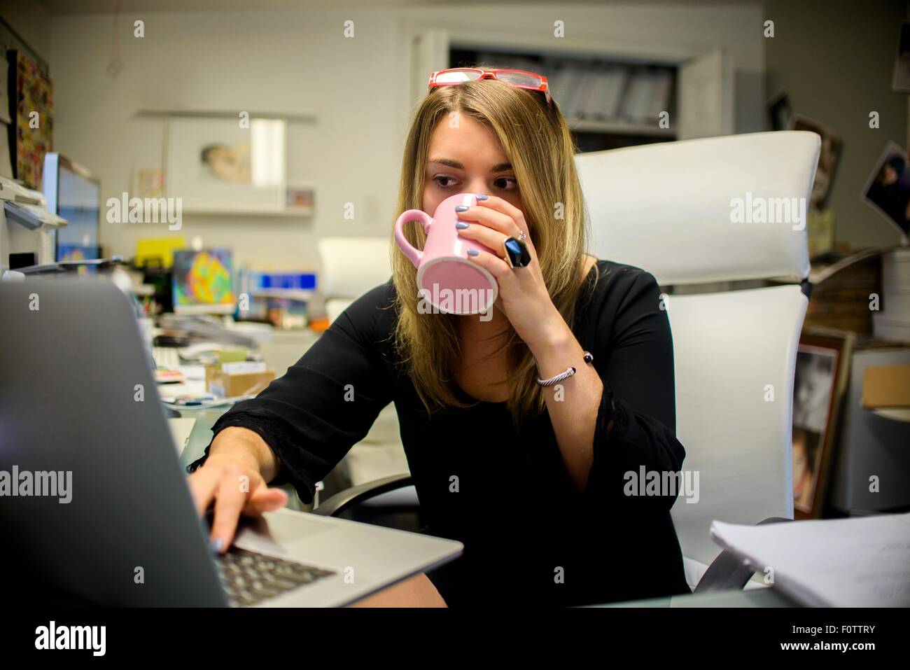 Young woman in office, sitting at desk, drinking coffee, using laptop Stock Photo