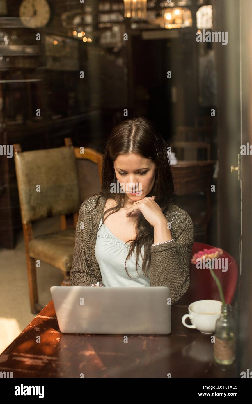 Mid adult woman sitting at table using laptop, hand on chin Stock Photo