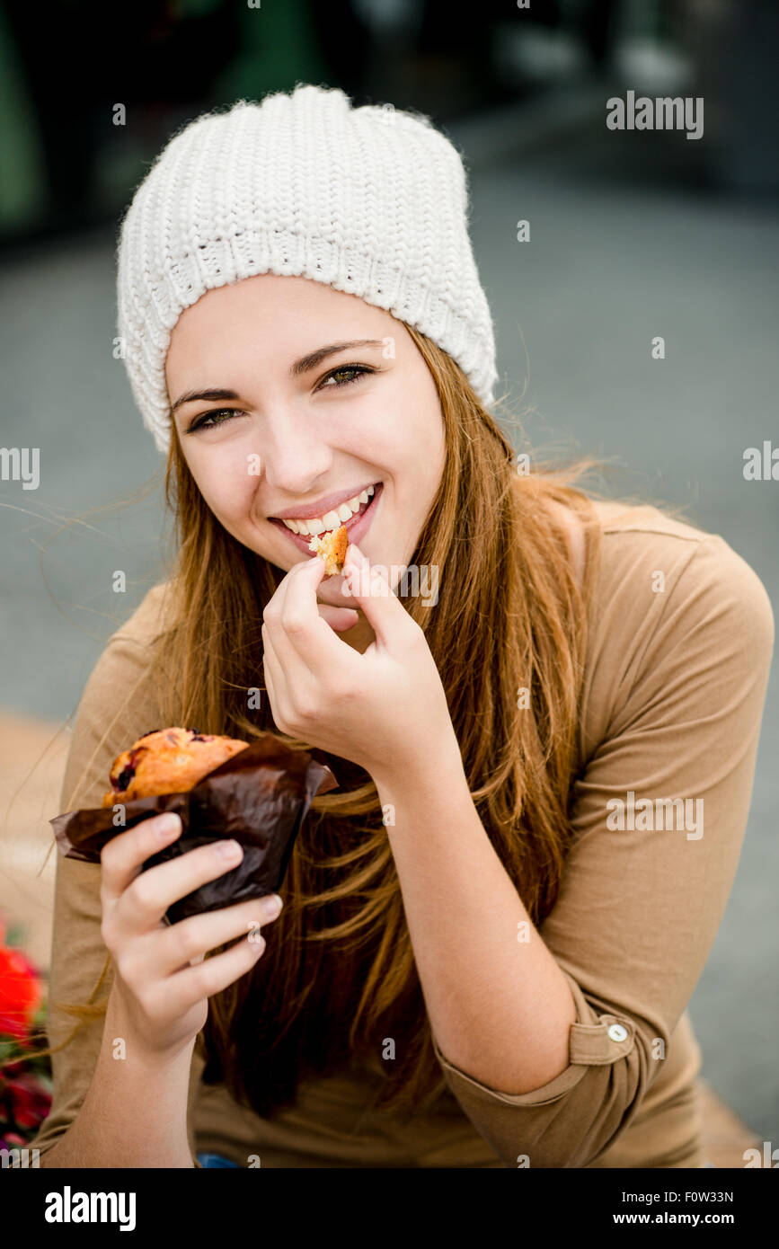 Young woman - teenager in cap eating muffin outdoor in street Stock Photo