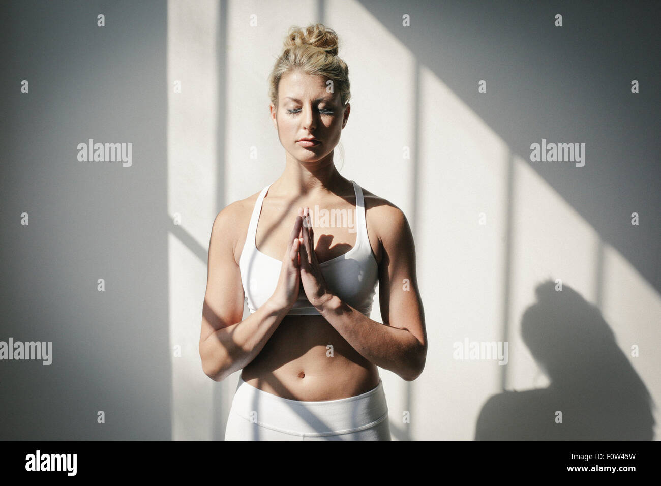 A blonde woman, her eyes closed, in a white crop top and leggings, standing in front of a white wall, doing yoga. Stock Photo