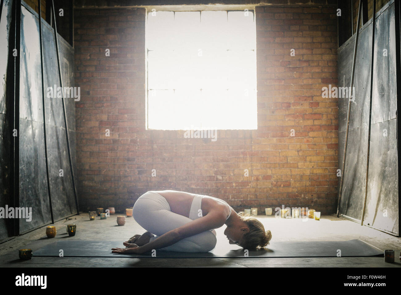 A blonde woman in a white crop top and leggings,kneeling on a mat on the floor surrounded by candles. Stock Photo