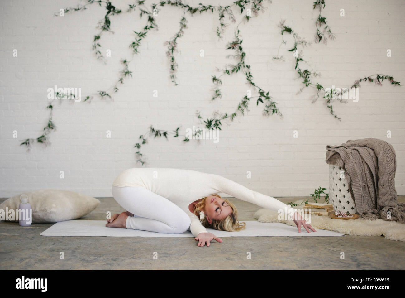 A blonde woman, in a white leotard and leggings, kneeling on a yoga mat with her arms stretched forwards. Stock Photo