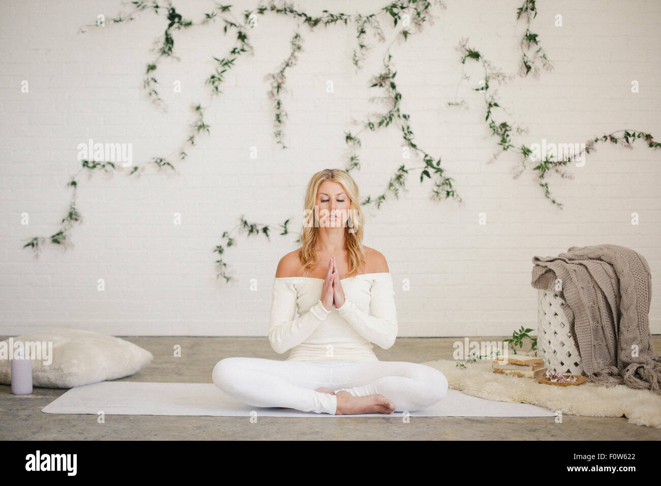 A blonde woman sitting on a white mat in a room, doing yoga. A creeper plant on the wall behind her. Stock Photo