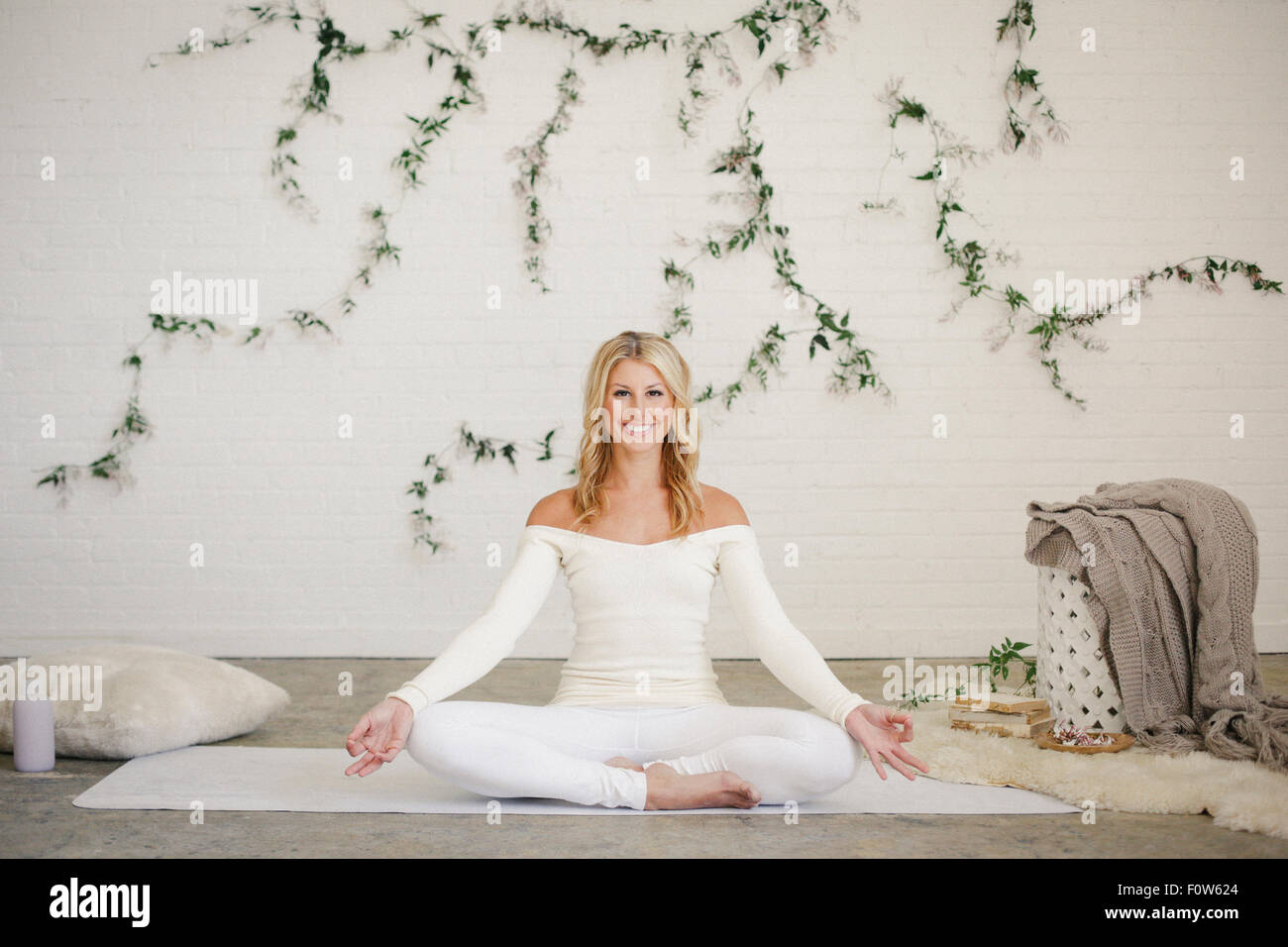 A blonde woman in a white leotard sitting on a white mat in a room, doing yoga. A creeper plant on the wall behind her. Stock Photo